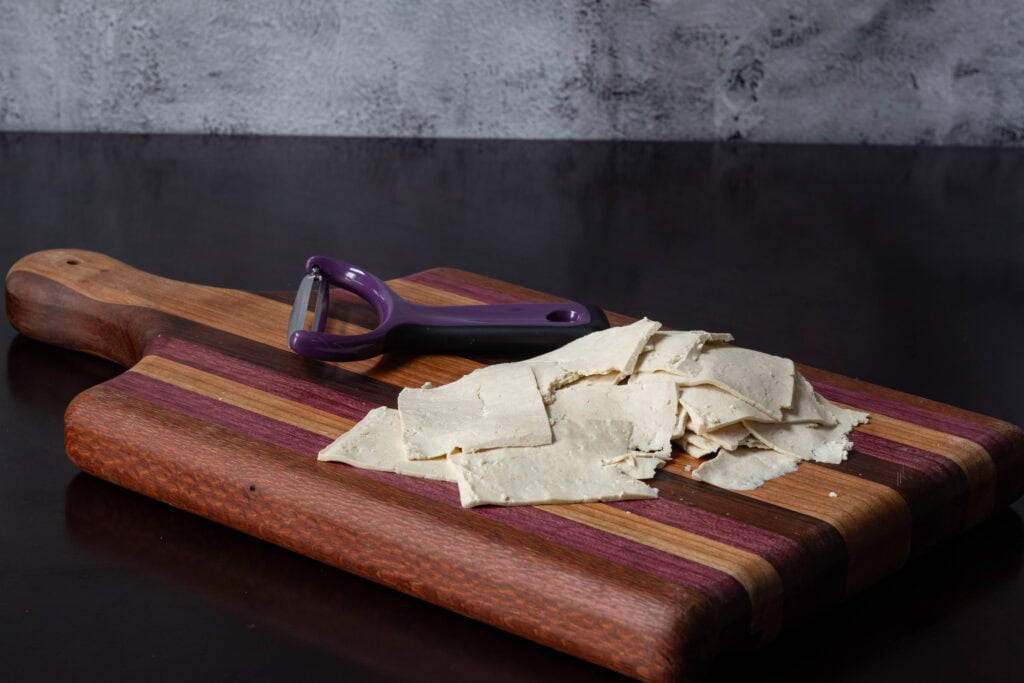 Wooden cutting board with thinly sliced tofu and wide vegetable peeler.