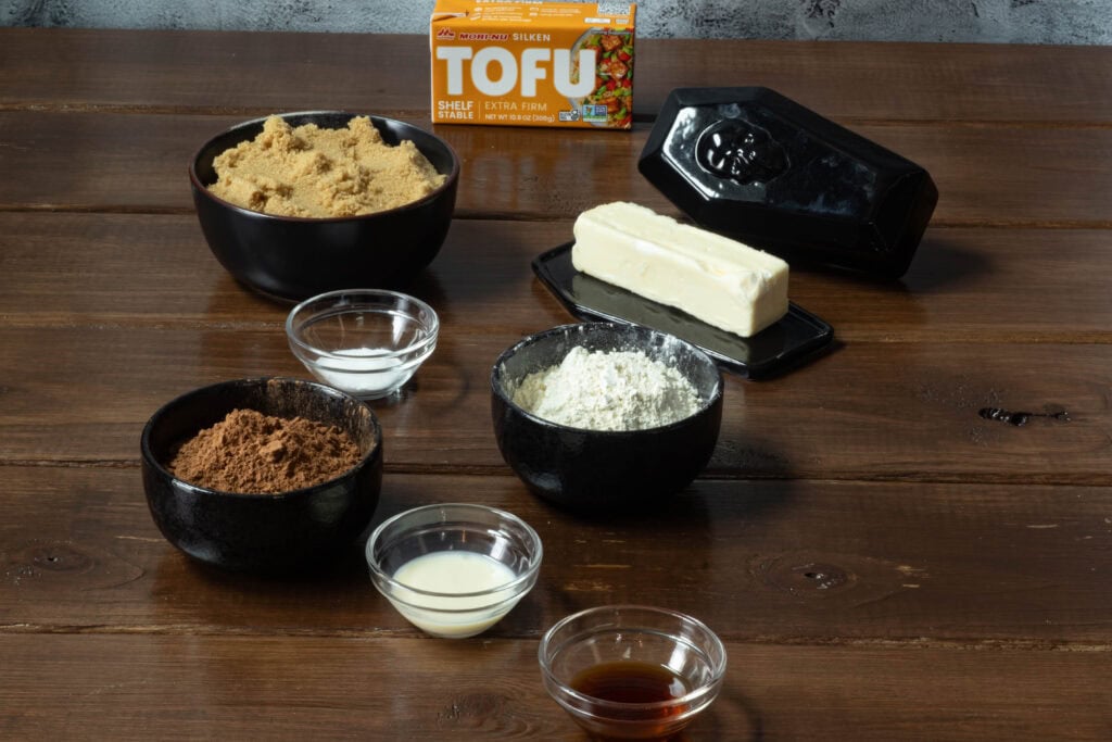 Protein brownie ingredients sitting on a wooden table - silken tofu, brown sugar, cocoa powder, soy flour, vegan butter, vanilla extract, salt, baking powder.
