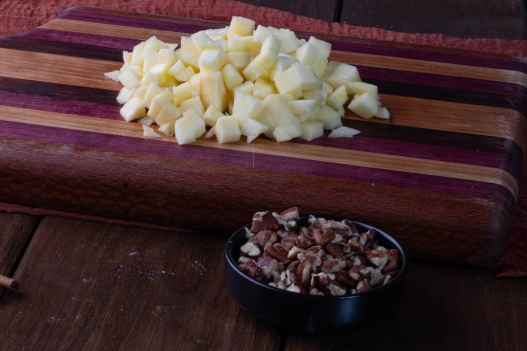 Chopped and peeled apples on a wooden cutting board next to a bowl of chopped pecans.