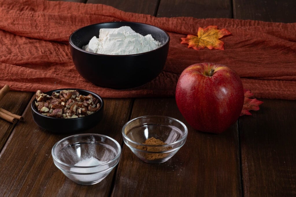 Pumpkin apple muffin ingredients siting on a table - chopped pecans, flour, apple, cinnamon, salt, baking powder and baking soda.