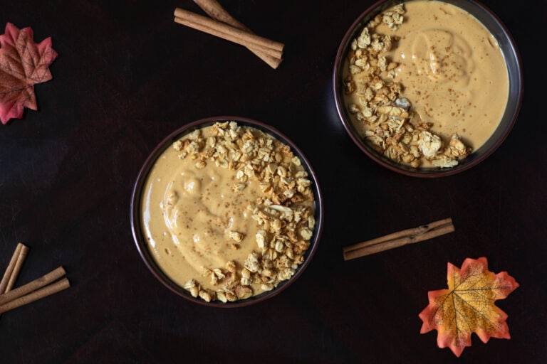 Two bowls with pumpkin pie smoothies topped with granola sitting on a wooden table. Scene is decorate with a fall vibe - leaves and cinnamon sticks.