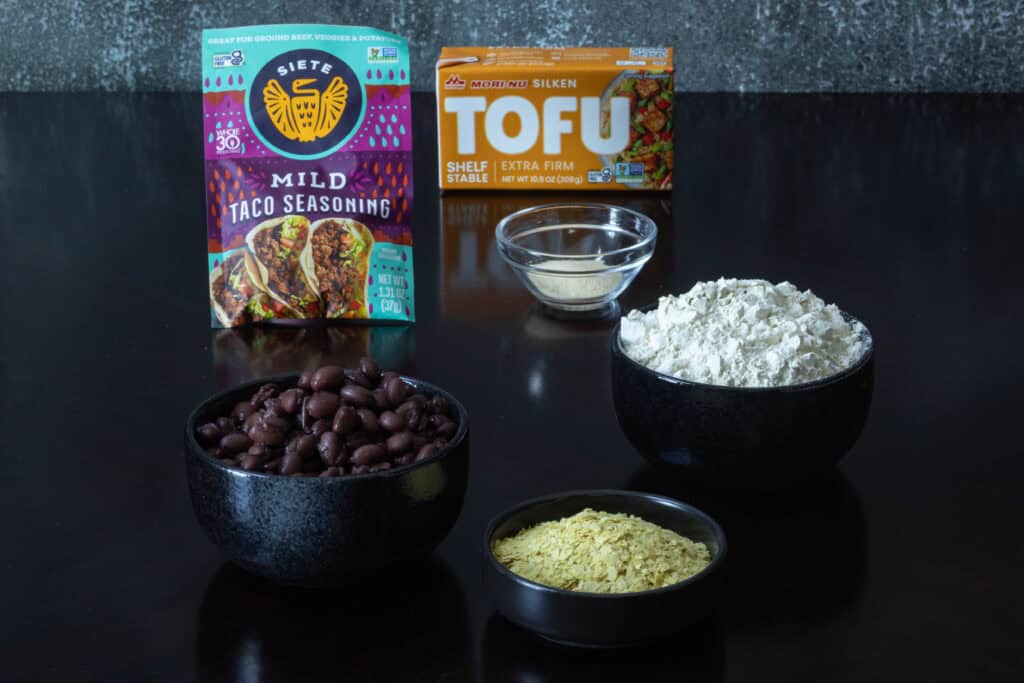 Seitan steak ingredients sitting on a wooden table - taco seasoning, other spices, silken tofu, vital wheat gluten, nutritional yeast and black beans.