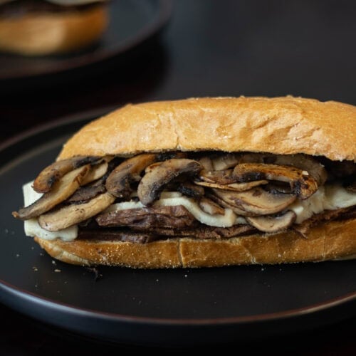 Plate with sandwich with shaved seitan steak, melted vegan mozzarella, and sauteed mushrooms.