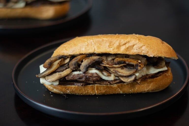 Plate with sandwich with shaved seitan steak, melted vegan mozzarella, and sauteed mushrooms.