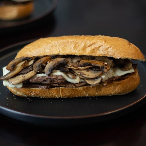 Plate with sandwich with shaved seitan steak, melted vegan mozzarella, and sauteed mushrooms.