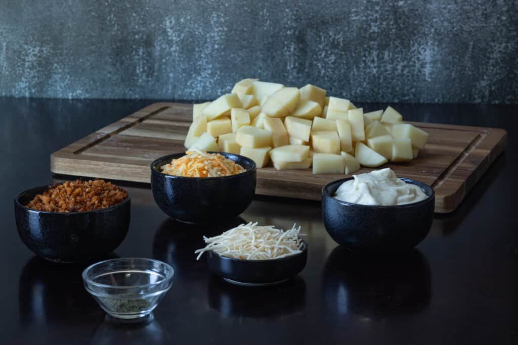 Twice baked potato casserole ingredients sitting on a wooden table - vegan bacon bits, dill, parmesan, shredded cheese, sour cream and peeled potato chunks.