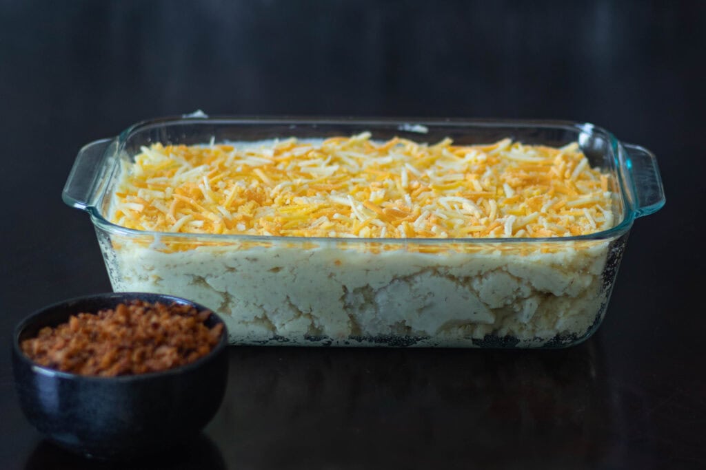 Large glass casserole dish with uncooked twice baked potato casserole, and a smaller bowl of vegan bacon bits sitting nearby.