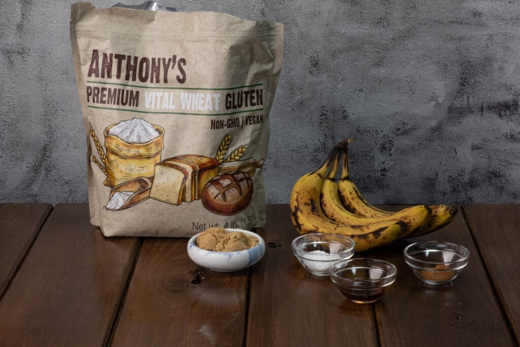 Banana bread seitan ingredients sitting on a table - vital wheat gluten, bananas, brown sugar, vanilla, salt, baking soda, baking powder and spices.