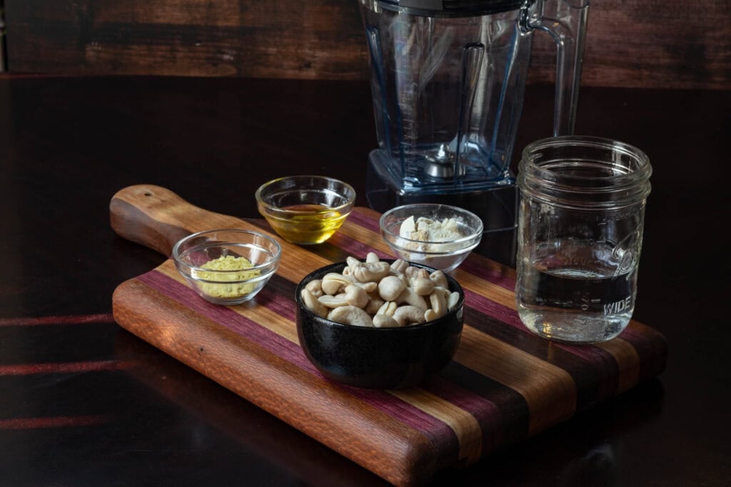 Ingredients for the liquid mozzarella sitting on a wooden cutting board - raw cashews, water, olive oil, nutritional yeast, spices, and a blender in the background.