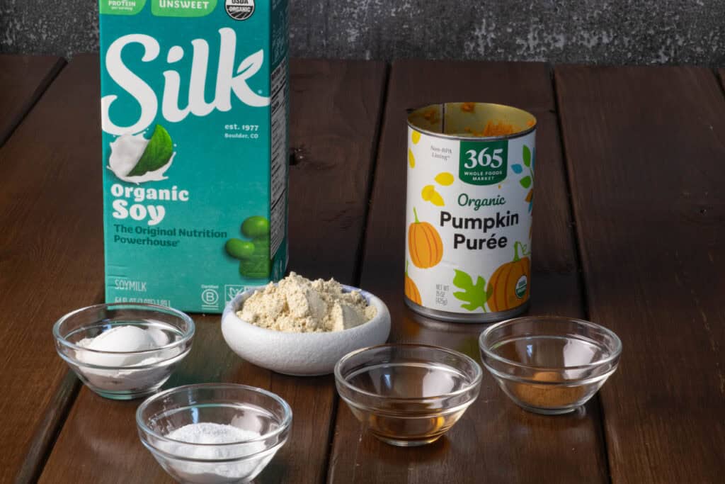 Protein pumpkin ingredients sitting on a wooden table - soy milk, pumpkin puree, protein powder, flour, baking powder, vanilla extract and cinnamon.