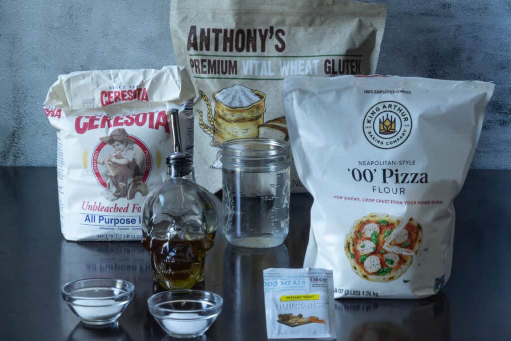 High Protein Pizza Dough ingredients sitting on a table - all-purpose flour, 00 flour, vital wheat gluten, water, olive oil, instant yeast, salt and sugar.