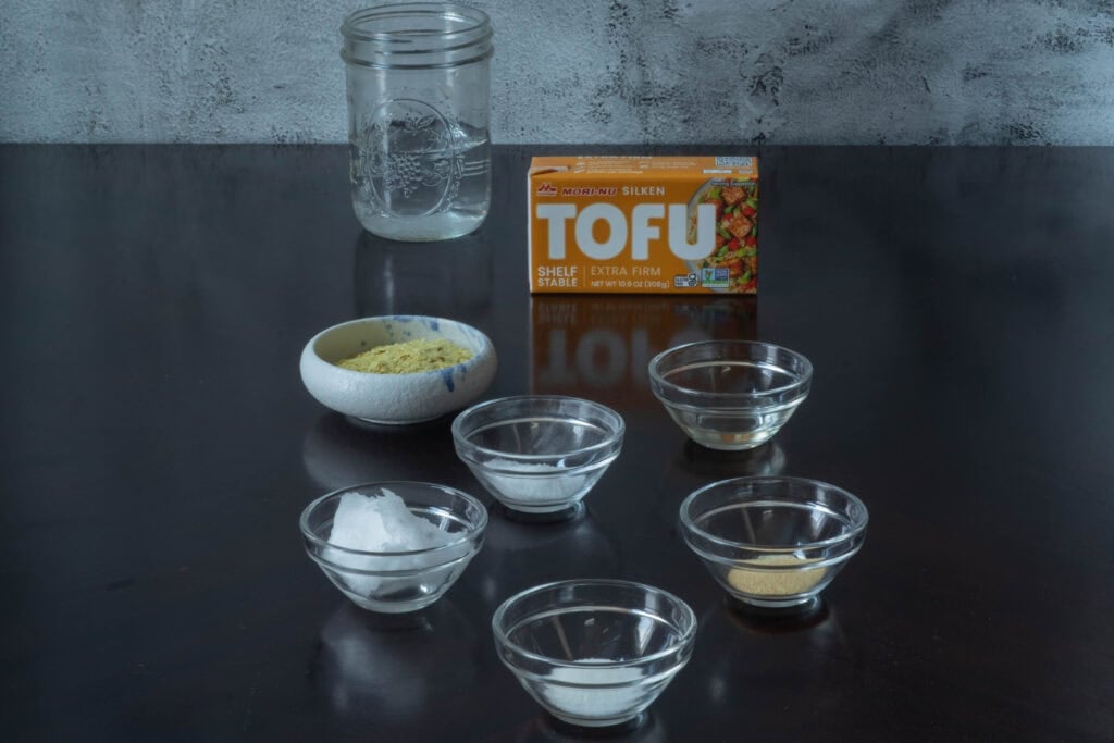 Stretchy soy mozzarella ingredients sitting on a table - water, silken tofu, nutritional yeast, tapioca flour, coconut oil, sugar, white whine vinegar and spices.