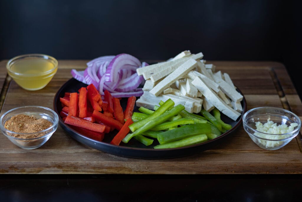 Tofu Fajita ingredients sitting on a wooden cutting board - veggies, cut tofu, taco seasoning, vegetable broth, and minced garlic.