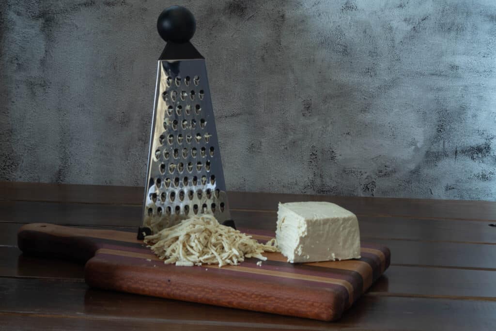 Vegetable grater, shredded tofu and half of a block of tofu sitting on a wooden cutting board.