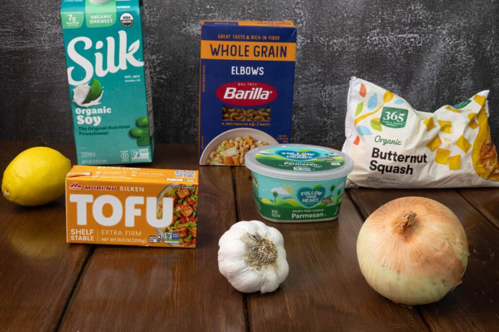 Ingredients sitting on a table - soy milk, silken tofu, lemon, garlic, onion, vegan parmesan, elbow macaroni, butternut squash.