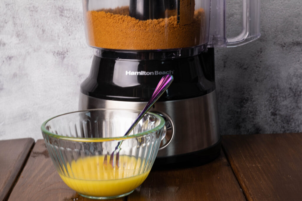 Crushed Biscoff cookies in a food processor and a glass bowl of melted butter.