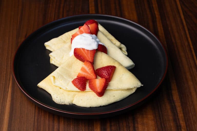 Vegan crepes on a black plate with strawberries and whipped cream.