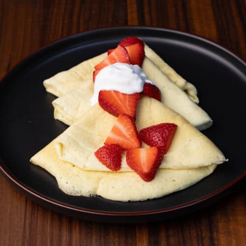 Vegan crepes on a black plate with strawberries and whipped cream.